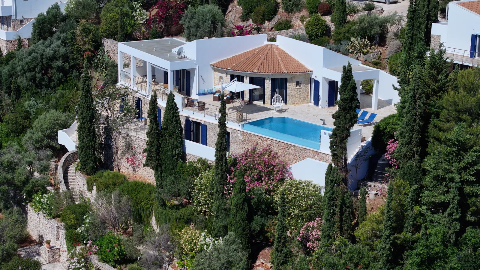 Aerial view of the first villa, its conical tiled roof and stone walls above a terrace with a rectangular pool, cypresses and flowering oleander on the slope below