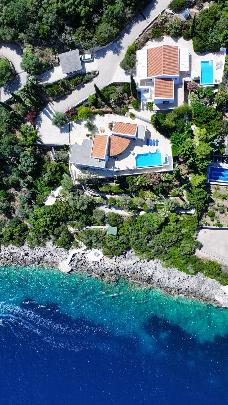 Top down aerial of the shoreline, sapphire and turquoise water with the white bathing platforms, both villas and twin pools among the gardens