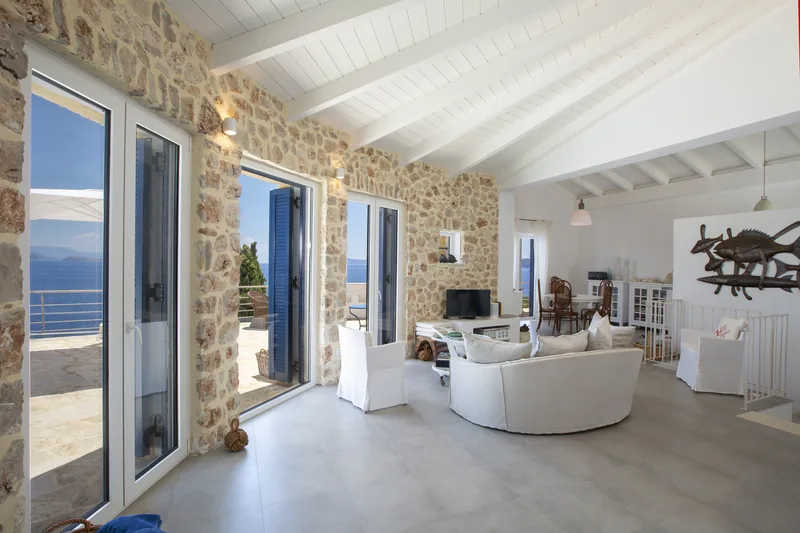 The main living room from its far end, rough stone walls and a white beamed ceiling, glazed doors opening to the terrace and the sea