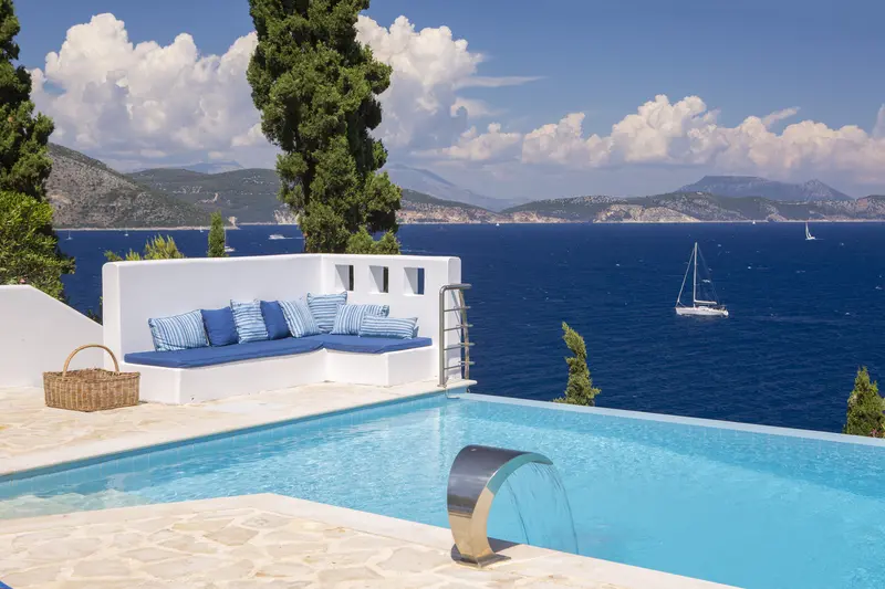 Corner of the infinity pool with white banquette and striped pillows, a lone sailboat on the strait beyond