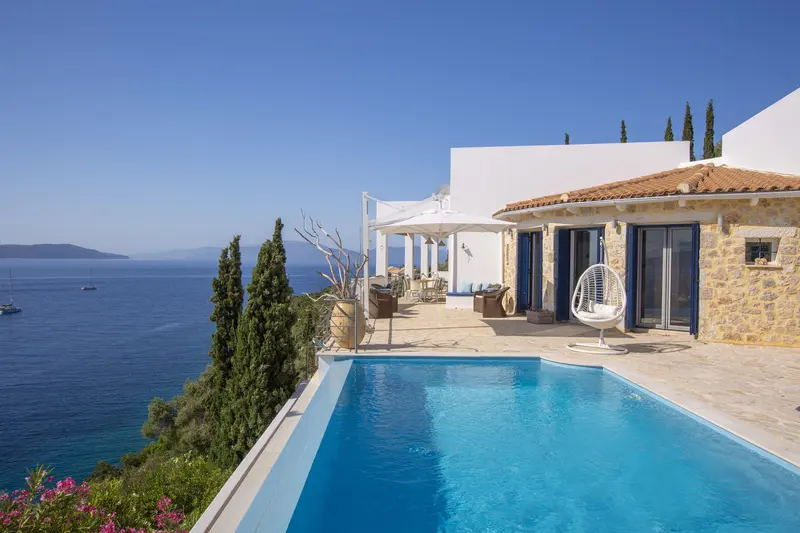 Stone and white villa with blue shutters above its infinity pool, cypress trees and the open Ionian Sea