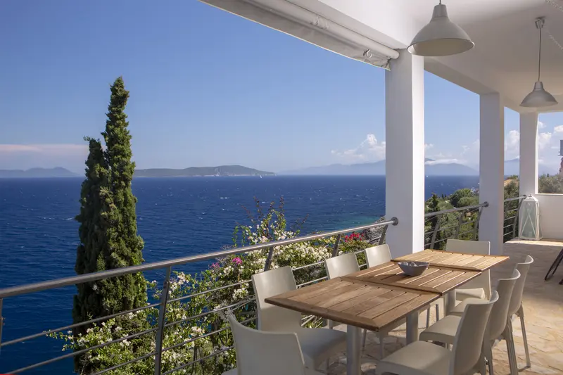 Covered dining terrace with a long teak table under an awning, cypress and white bougainvillea framing the sea