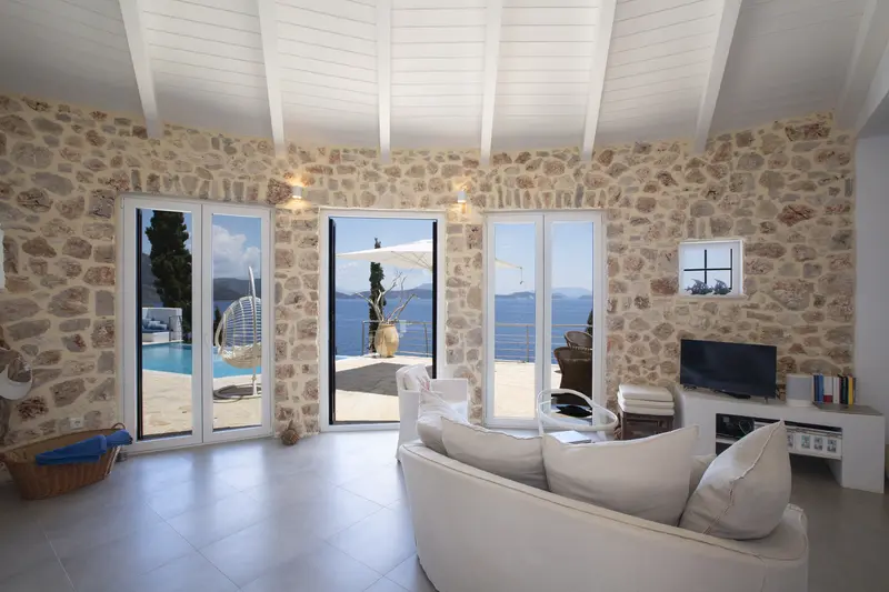 Living room with exposed stone wall and white beamed cathedral ceiling, linen sofa and French doors framing the pool and sea