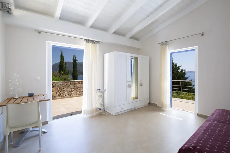 Airy white bedroom with curtained glass doors to a broad stone walled terrace and views over cypress to the sea